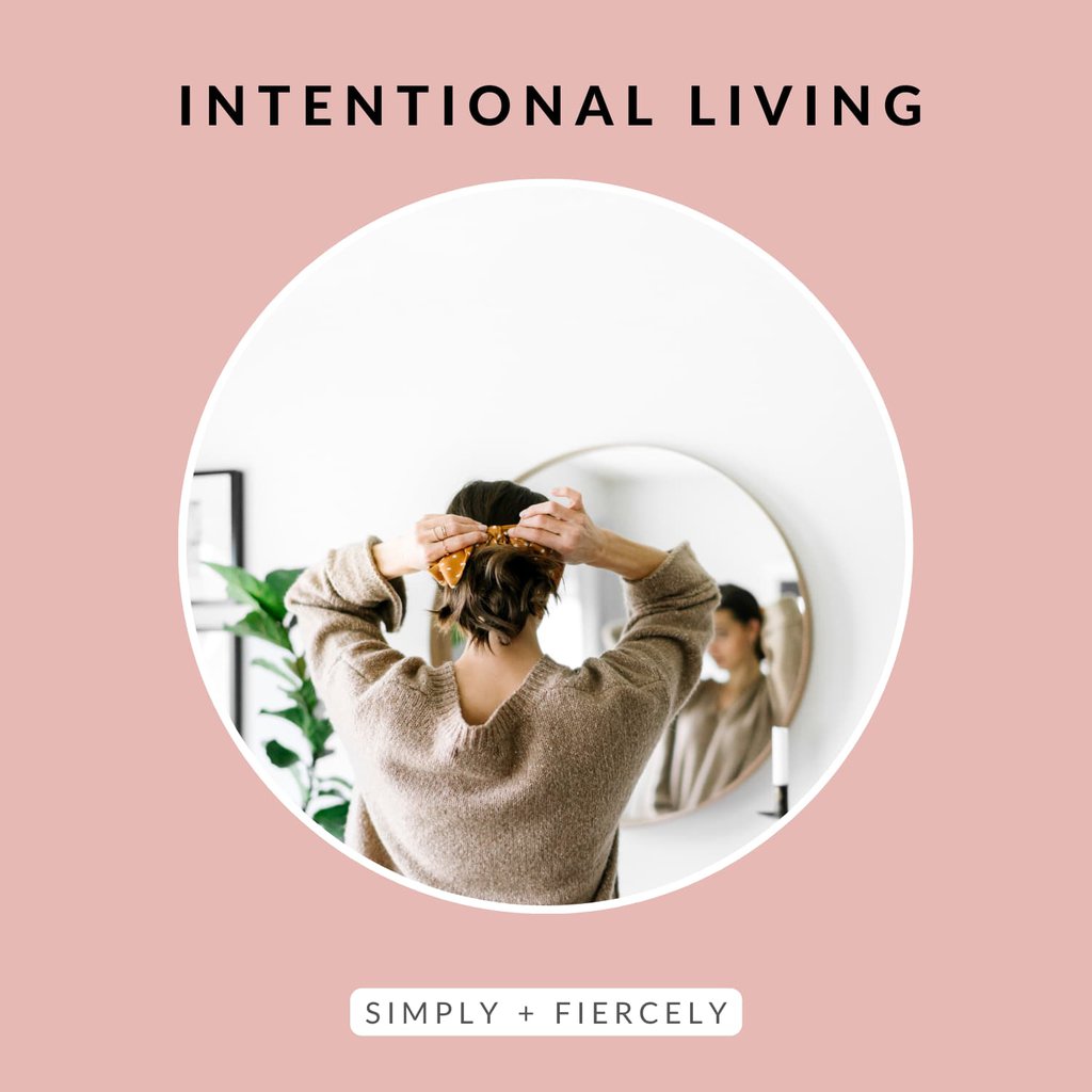 A pink circle frame with the text “intentional living” at the top and the text “Simply + Fiercely” at the bottom. In the frame is a woman looking in the mirror while tying up her dark hair. You can’t see her face.