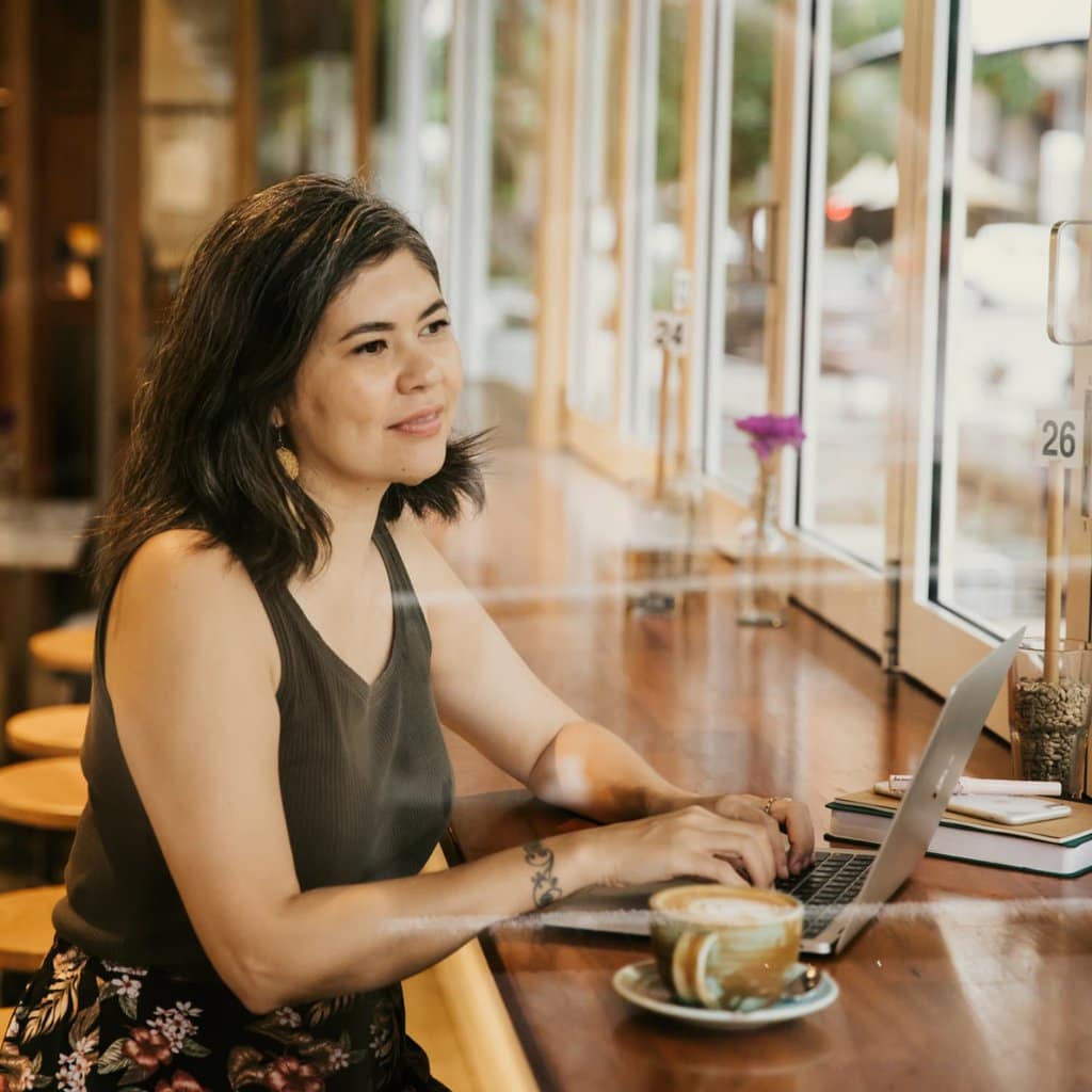 A dark haired woman on a laptop in a cafe looking out the window.