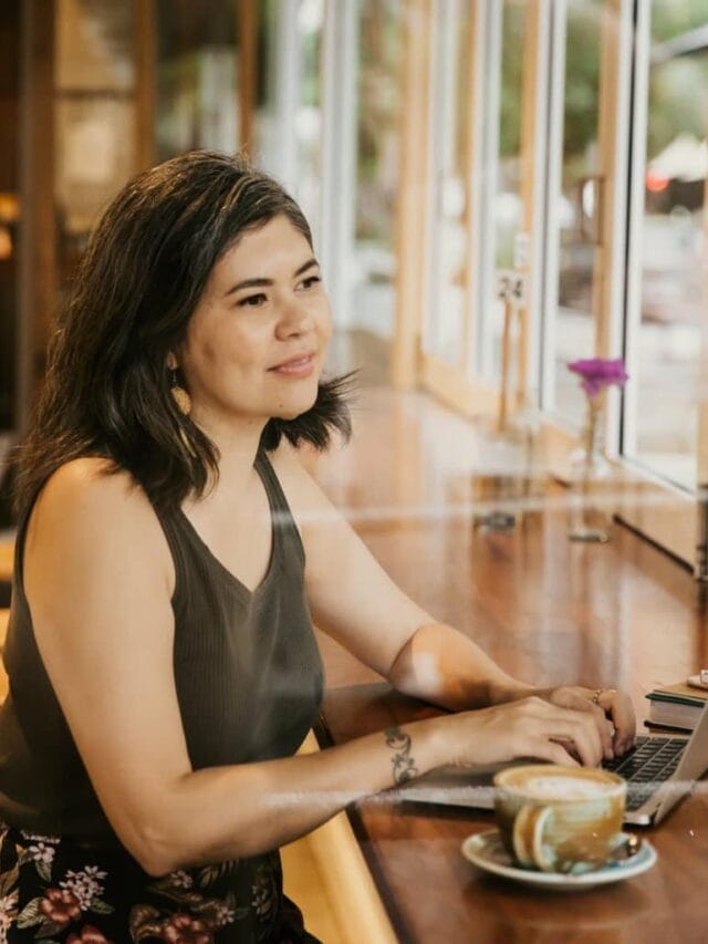 A dark haired woman on a laptop in a cafe looking out the window.