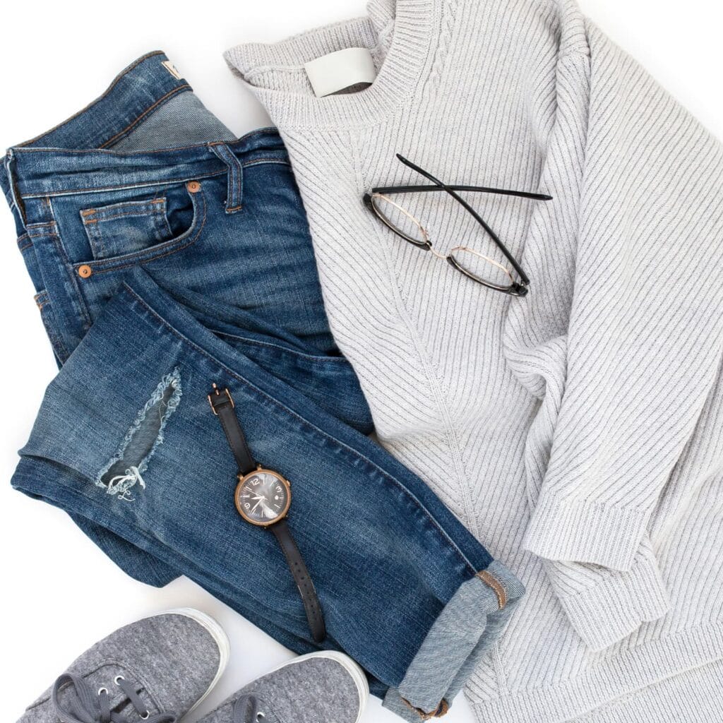 Flat lay with grey sweater, blue jeans, grey shoes, a watch and glasses, against a white background
