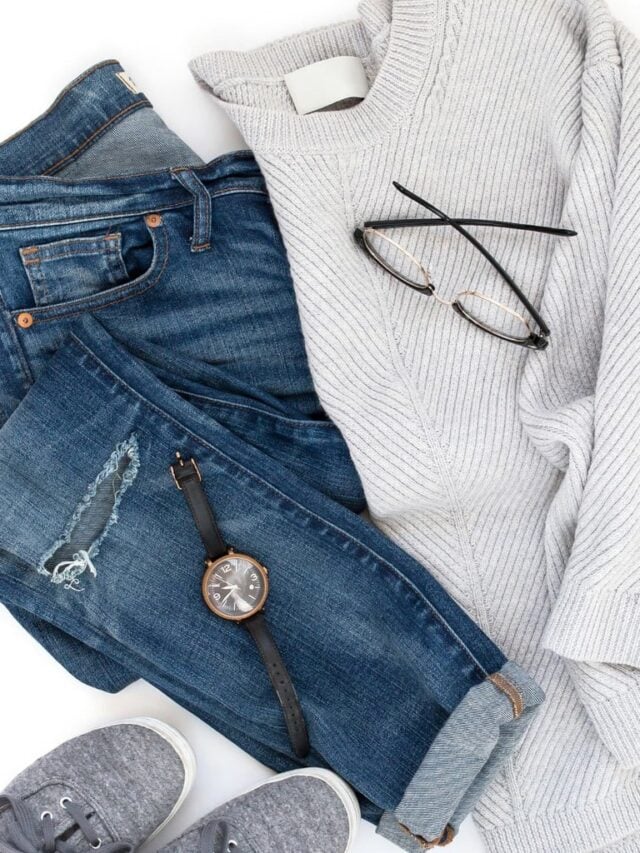 Flat lay with grey sweater, blue jeans, grey shoes, a watch and glasses, against a white background