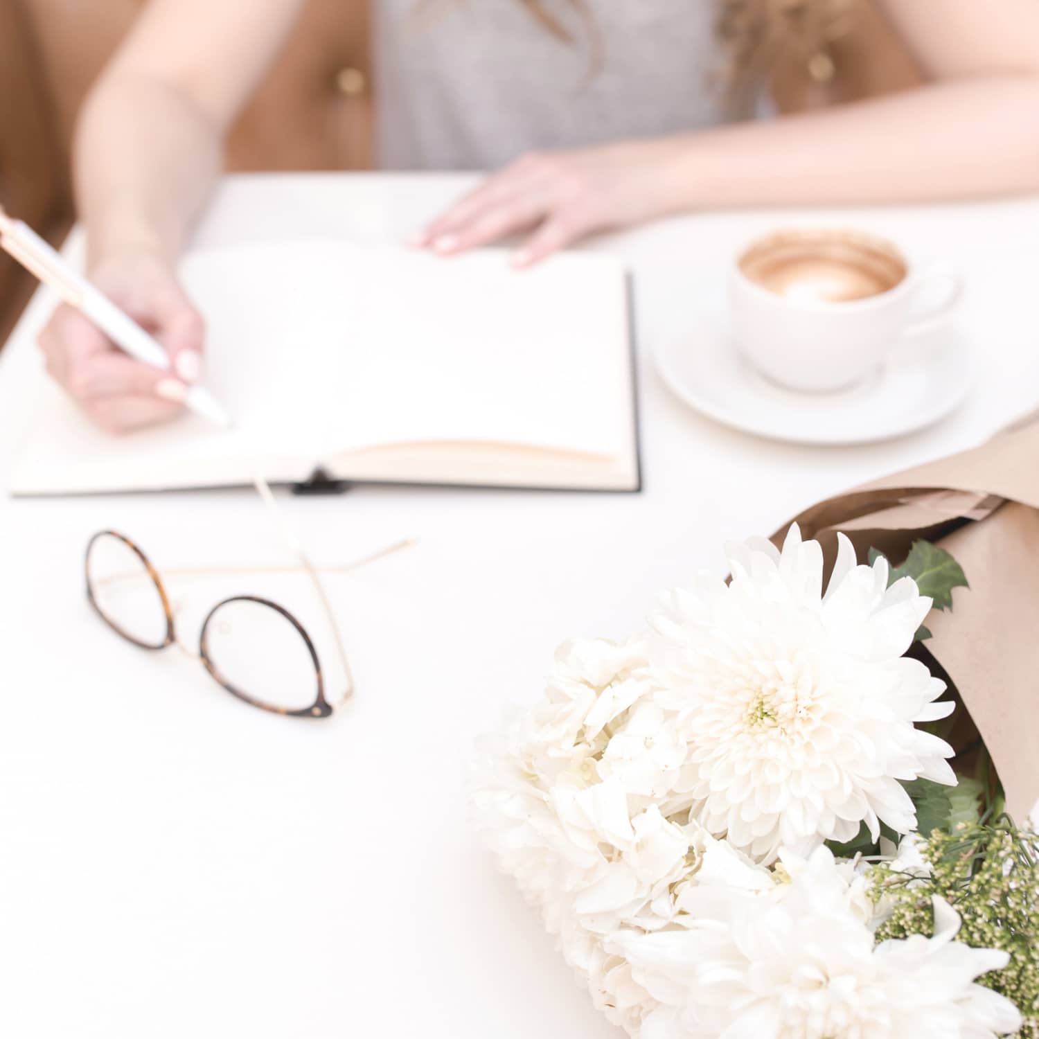 An image of a woman writing in a journal with a cup of coffee, a pair of glasses and a bouquet of white flowers laying on the table beside her.