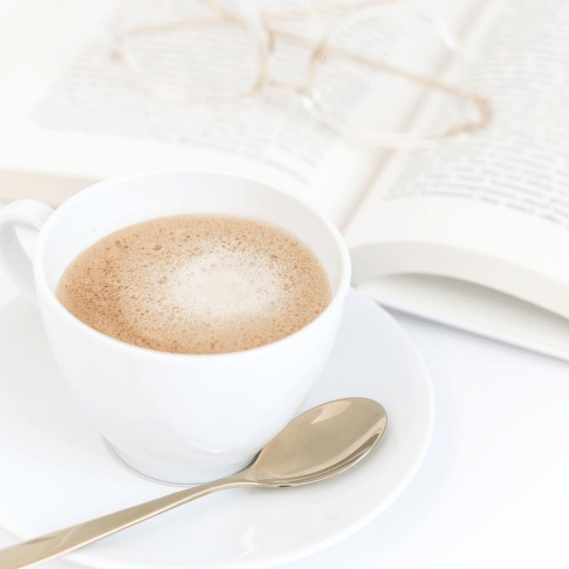 A book with eyeglasses and cup of coffee on saucer with tea spoon.