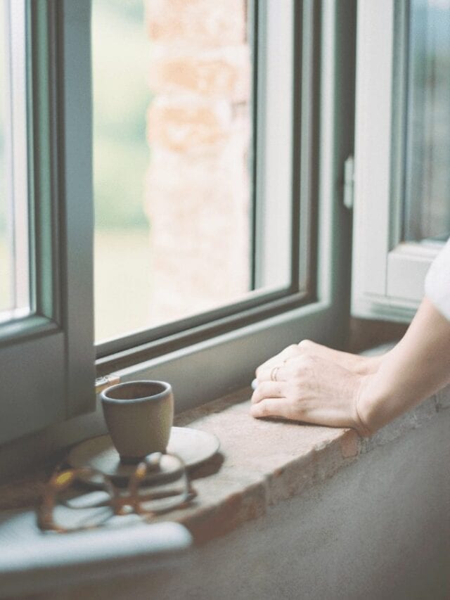 A woman looking out a window with eye glasses and a coffee mug beside her.