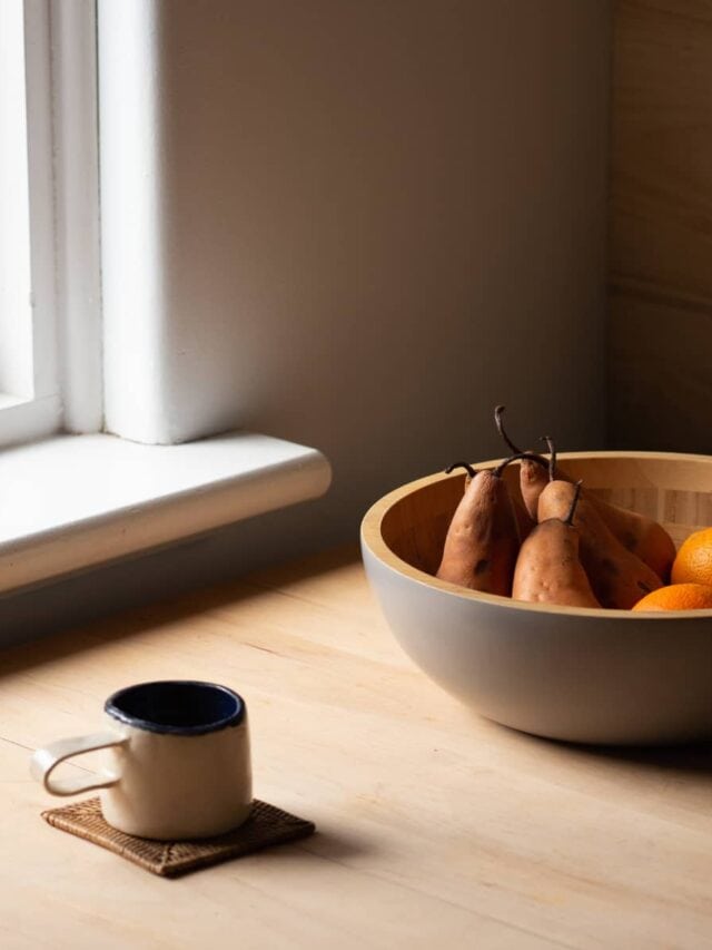 A mug and bowl of fruit sitting on a table in front of a window.