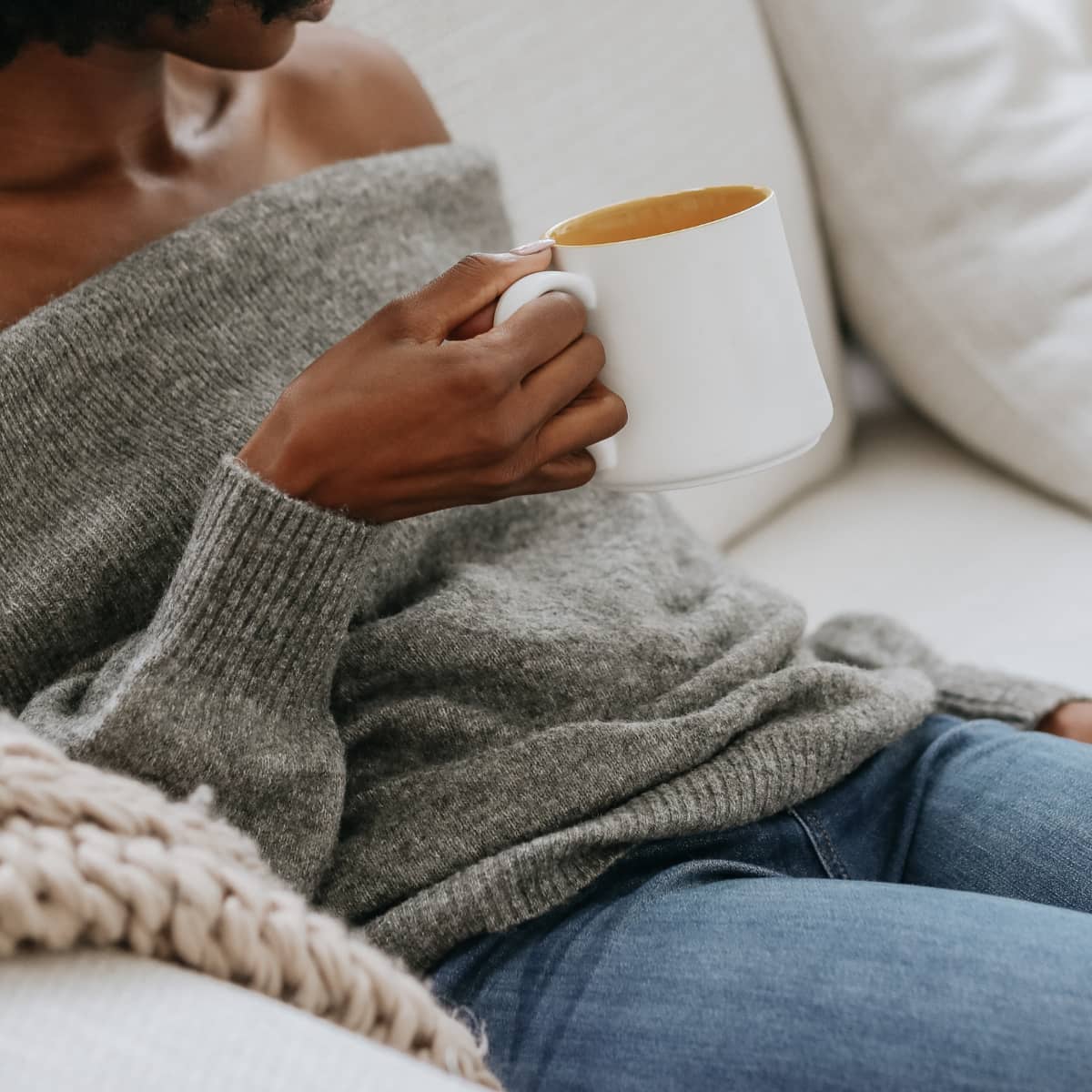 A woman wearing a grey sweater and jeans sitting on a sofa holding a cup of coffee