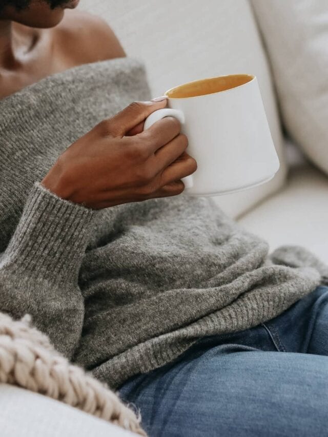 A woman wearing a grey sweater and jeans sitting on a sofa holding a cup of coffee
