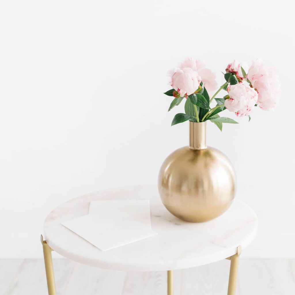 A side table with a gold vase holding pink peonies and a note pad on it
