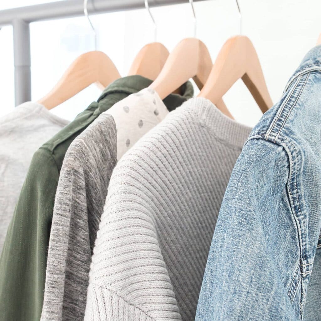 A minimalist closet with sweaters and a denim jacket hanging on wooden hangers.