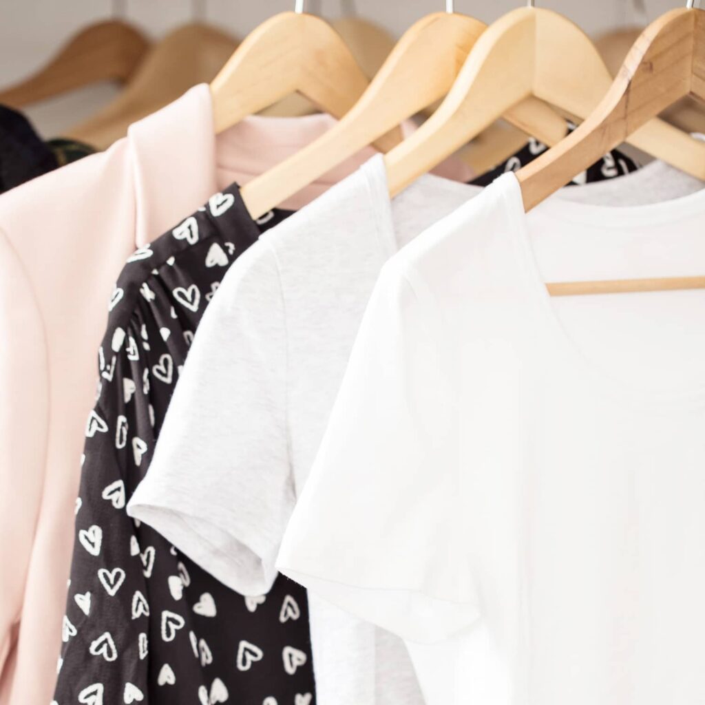 a white box with a pink jacket, a black top with white hearts, and 2 white t-shirts hanging on wooden hangers in the background.