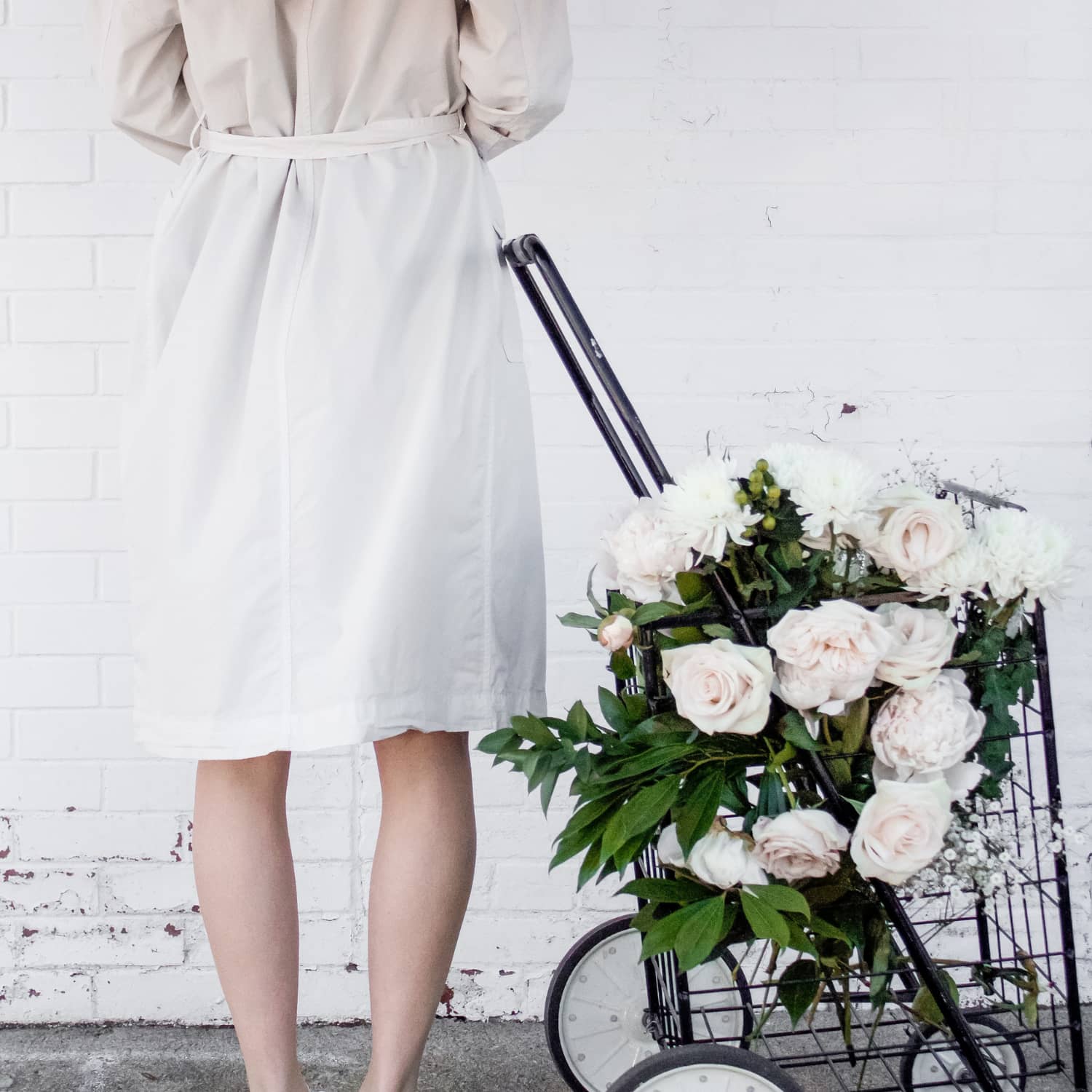 A woman facing away from the camera wearing a light trench coat standing beside a basket of flowers against a brick wall