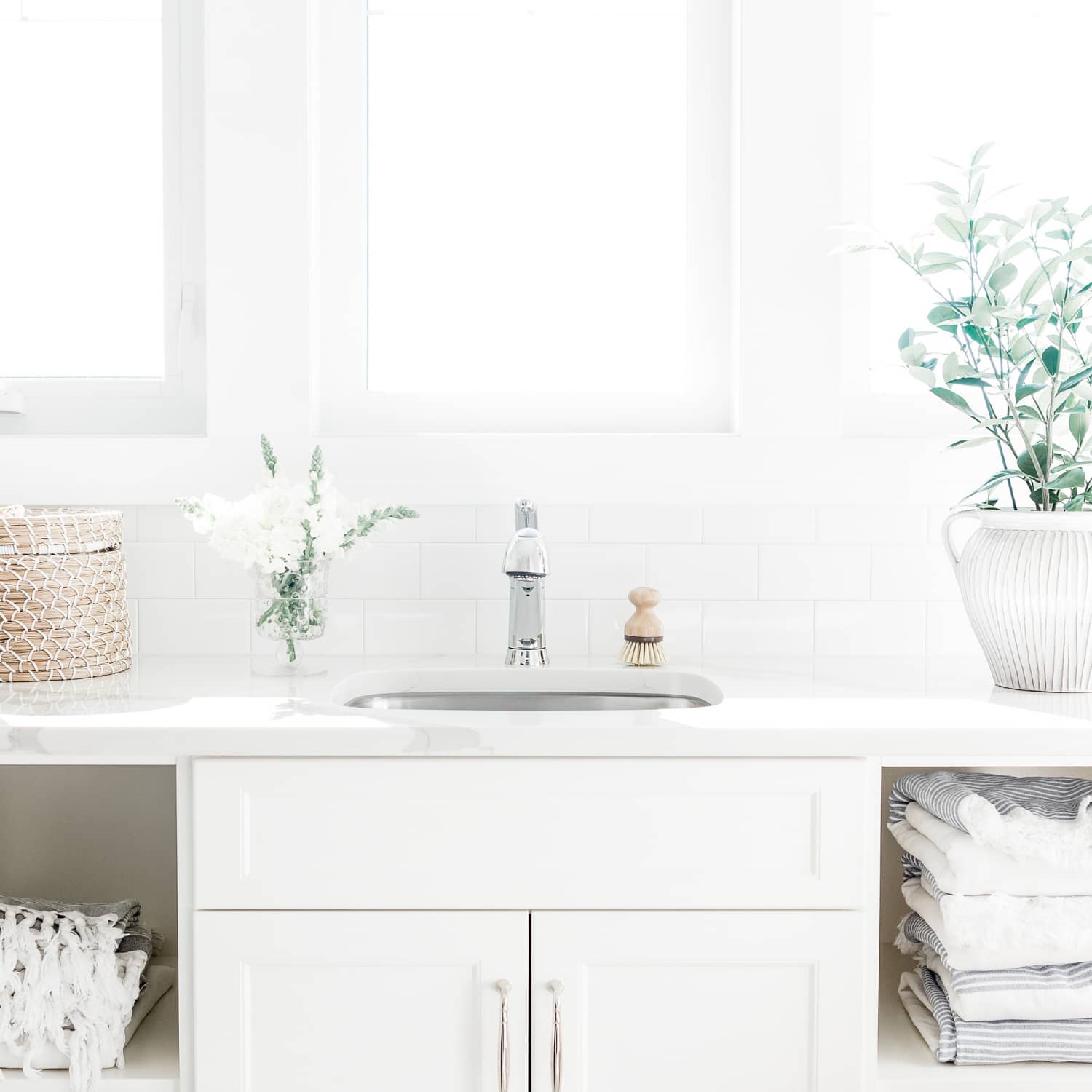 A minimalist laundry room with white cabinets, folded grey striped linens, a small basket, and a plant against a white background.