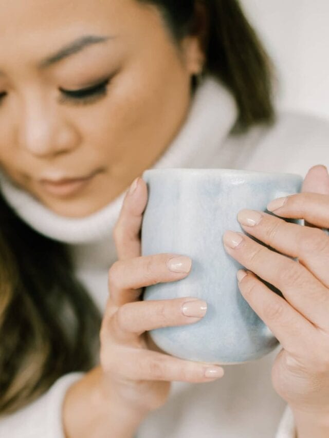 A closeup of a woman wearing a cream turtleneck and holding a light blue mug while looking down.