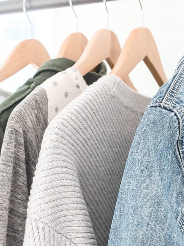 A minimalist closet with sweaters and a denim jacket hanging on wooden hangers.