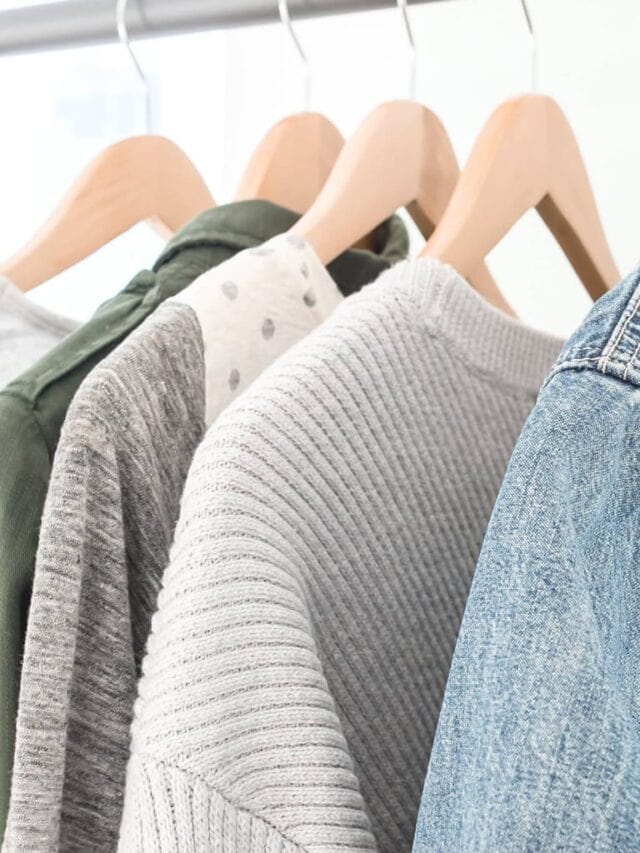 A minimalist closet with sweaters and a denim jacket hanging on wooden hangers.
