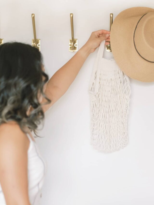 A woman reaching for a tan coloured hat that's hanging on a wall hook.