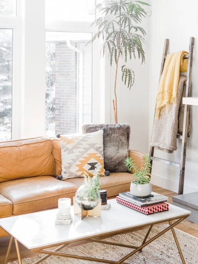 A minimalist living room with a mustard colored sofa with throw pillows, a coffee table with a stack of books, plants, and candles on it. A large plant is in front of large windows and a wooden blanket ladder is leaning against the wall.