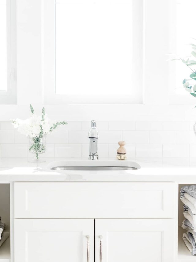 A minimalist laundry room with white cabinets, folded grey striped linens, a small basket, and a plant against a white background.