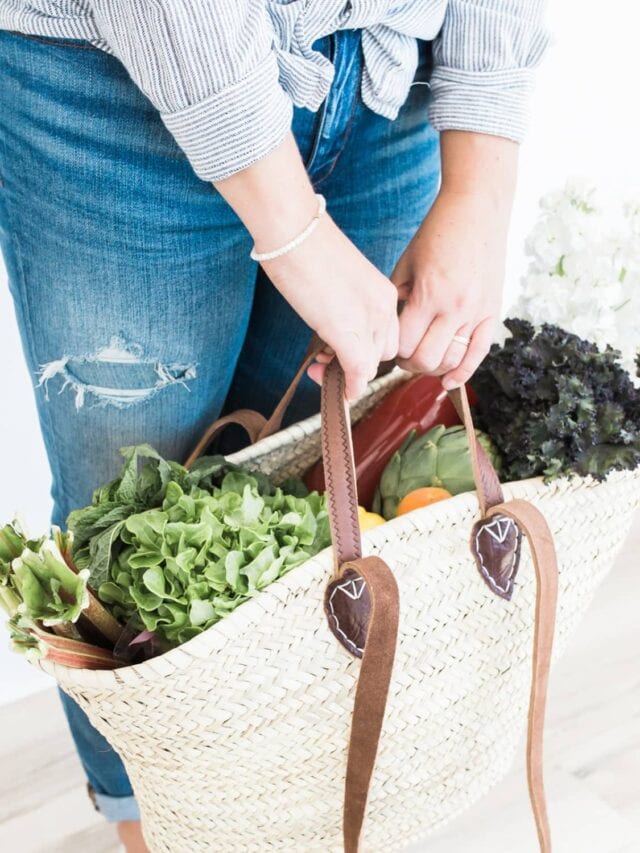 A woman wearing blue jeans and a stripped button up top holding a wicker bag full of fresh vegetables.