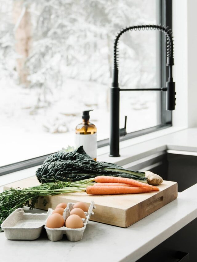 A minimalist kitchen with black cupboards, here is a wooden cutting board with carrots, kale, and ginger on a white countertop, a carton of brown eggs, a sink, and a black faucet all in front of a large window.