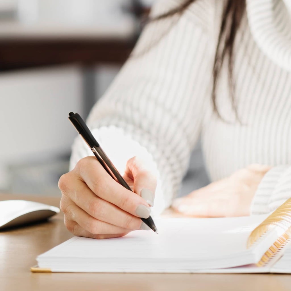 "7 Powerful Benefits of Journaling Why I RecommendA woman wearing a white turtleneck writing in an open coil-bound journal.