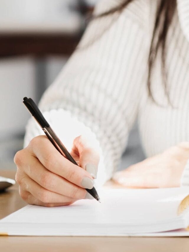 "7 Powerful Benefits of Journaling Why I RecommendA woman wearing a white turtleneck writing in an open coil-bound journal.