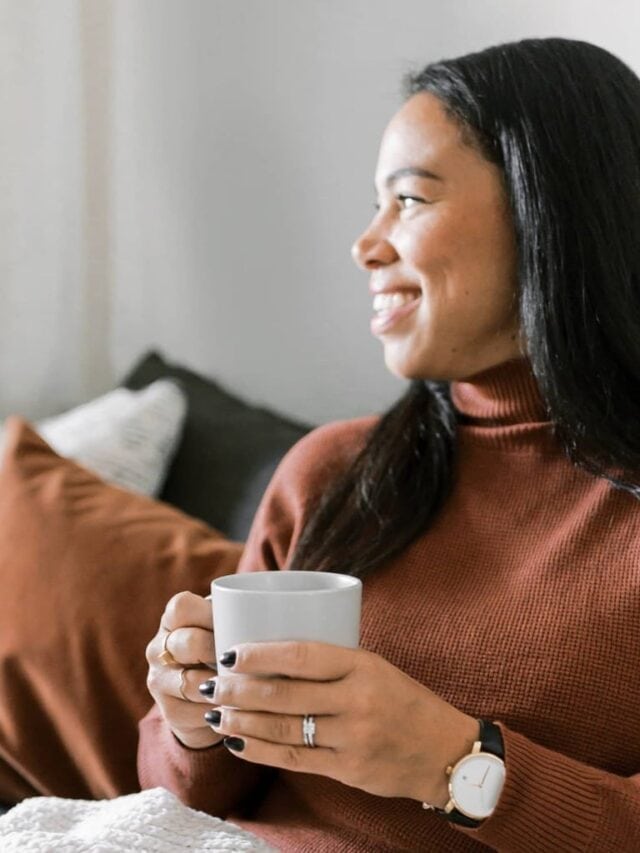 A smiling woman wearing a taupe turtleneck holding a cup of coffee sitting on a sofa.