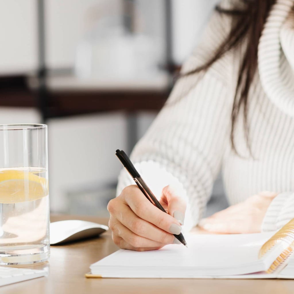 A woman writing in a journal with a glass of lemon water on the table beside her.