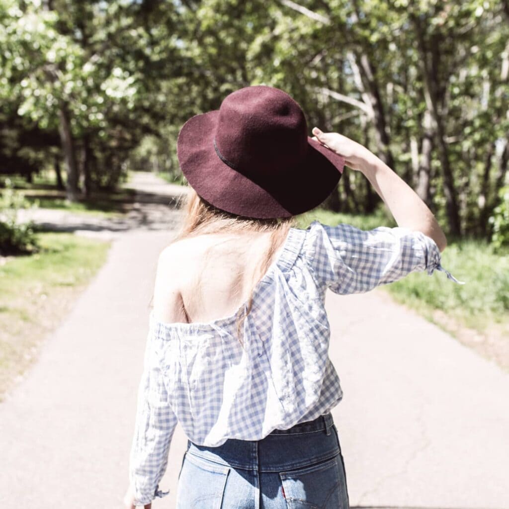 A woman wearing blue jeans, a blue checkered top, and a burgundy felt hat walking on a sidewalk outside towards some trees.