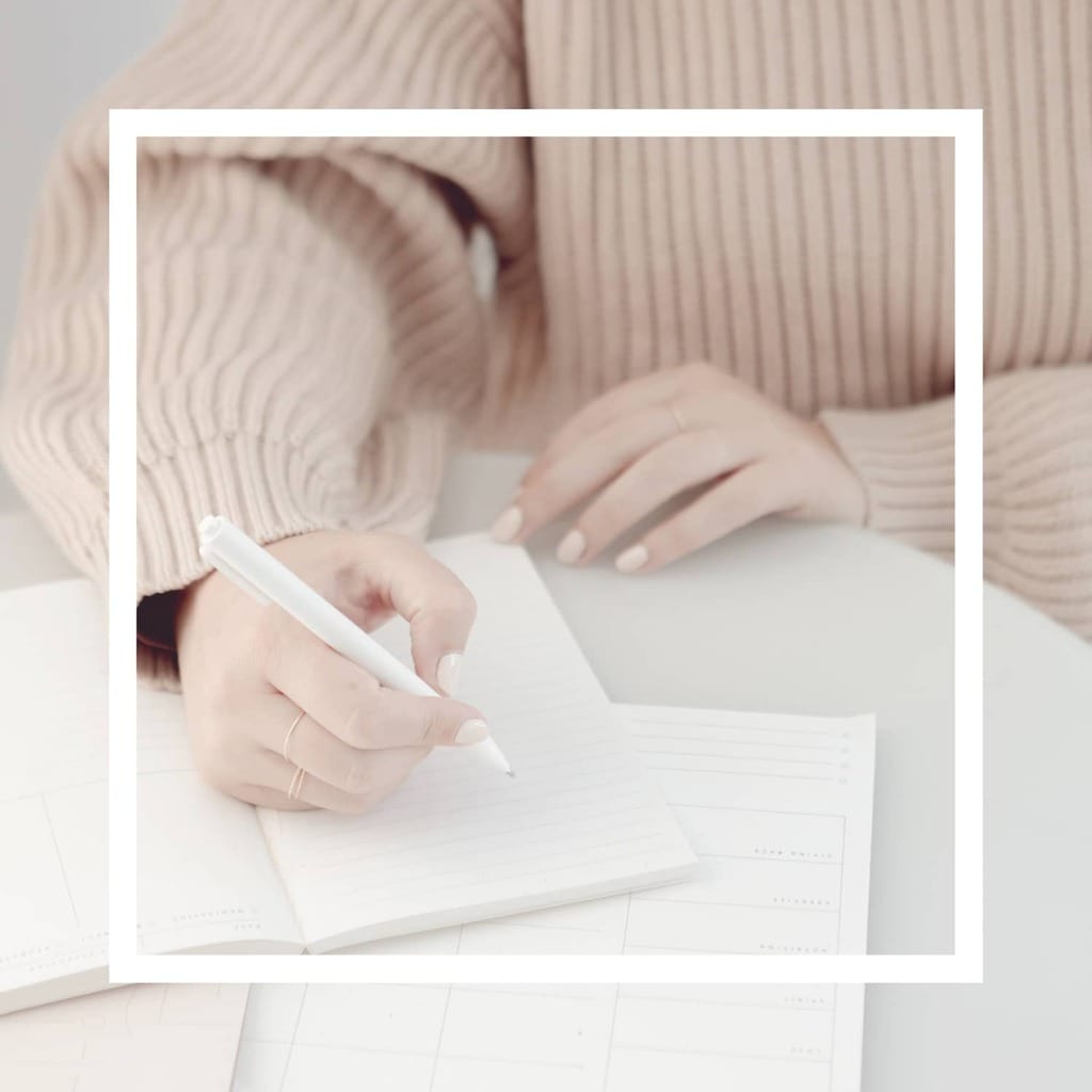 A white square overlay with an image of a woman wearing a pale pink sweater holding a white pen writing in a journal in the background.,