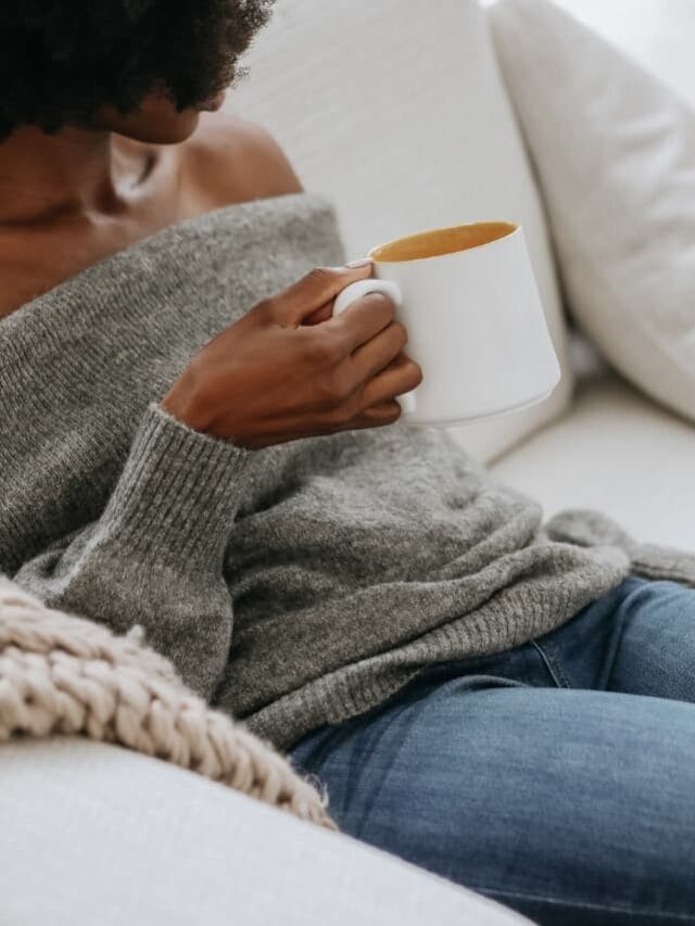 A woman sitting on a sofa, wearing a grey sweater and holding a grey coffee mug