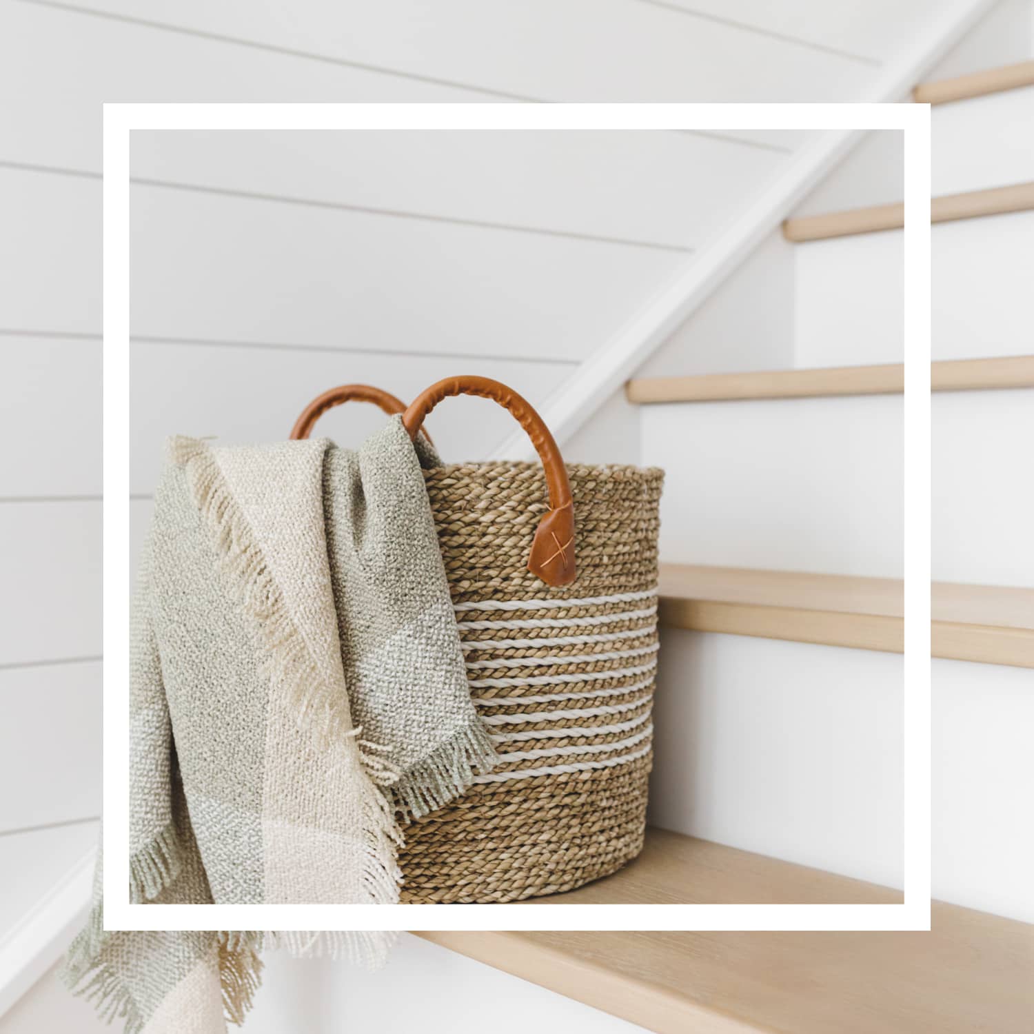 A straw basket with leather handles holding a beige plaid blanket sitting on a wooden staircase with a white frame overlay