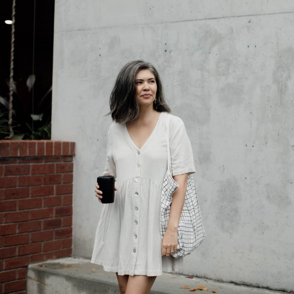 A woman wearing a white dress looking away from the camera, she's carrying a white and black checkered bag and holding a coffee standing in front of a concrete wall with a brick partition to the side.