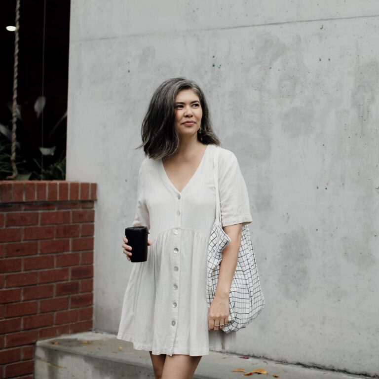 A woman looking away from the camera wearing a white dress carrying a white and black checkered bag and holding a coffee standing in front of a concrete wall with a brick partition to the side.