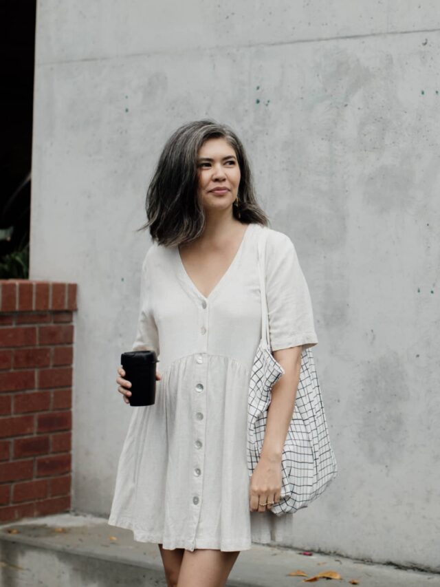 A woman looking away from the camera wearing a white dress carrying a white and black checkered bag and holding a coffee standing in front of a concrete wall with a brick partition to the side.
