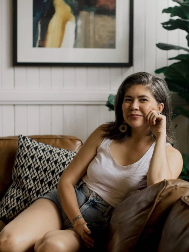 A woman wearing denim shorts and a white tank top sitting on a brown leather sofa and smiling into the camera in the background.