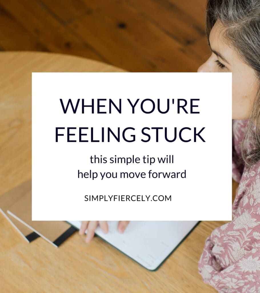 "When You Feel Stuck In Life, This Tip Will Help You Move Forward" in a white box with a woman wearing a purple and white floral top sitting at a wooden table holding a pen and a journal while looking away from the camera in the background.