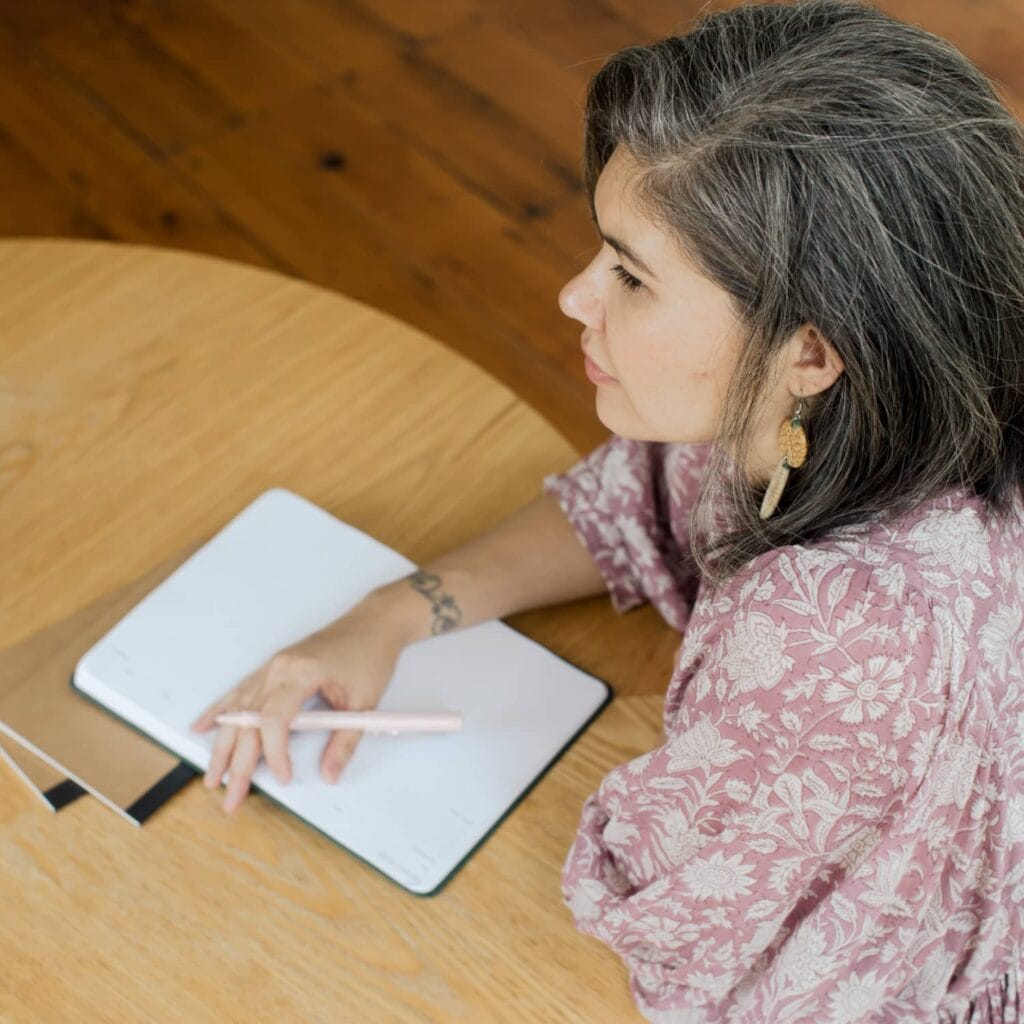 A woman wearing a purple and white floral top sitting at a wooden table holding a pen and a journal while looking away from the camera