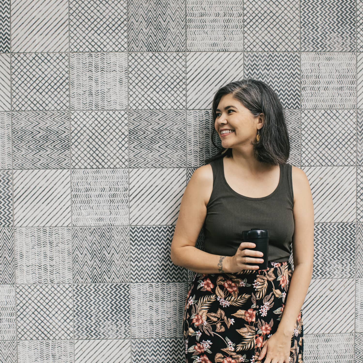A smiling woman wearing a black tank top and floral skirt standing in front of a grey and white patterned wall.