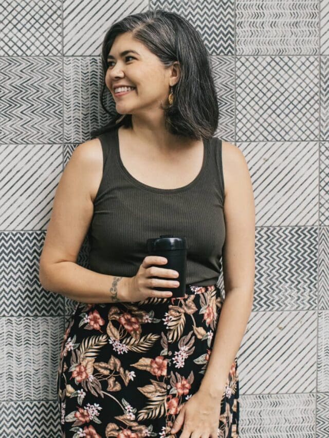 A smiling woman wearing a black tank top and floral skirt standing in front of a grey and white patterned wall.