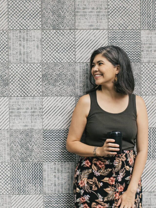 A smiling woman wearing a black tank top and floral skirt standing in front of a grey and white patterned wall.