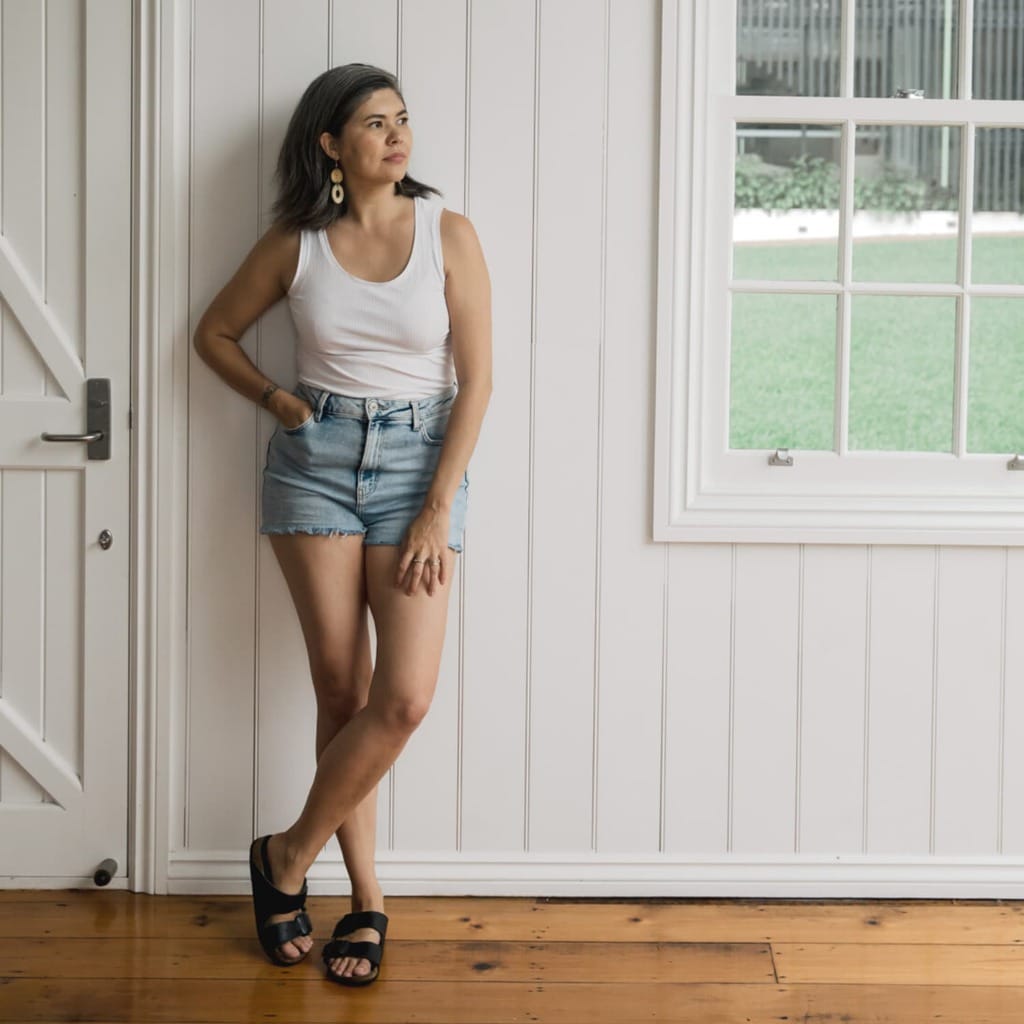 A woman wearing a white tank top, denim shorts, and sandals leaning against a white wooden wall looking towards a window