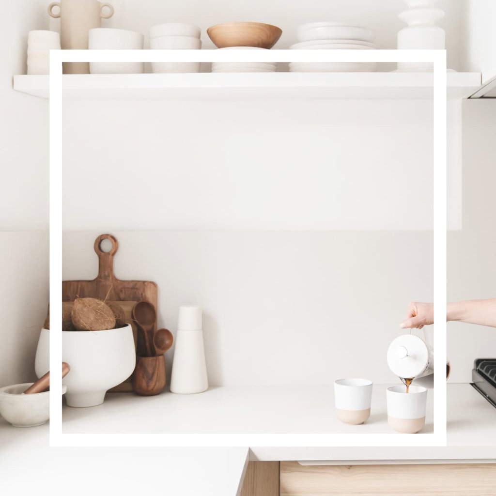 A minimalist kitchen with stacked white bowls on a white shelf. On the counter there's a wooden cutting board, wooden spoons in a bowl, more white bowls, and a hand coming from the right of the image pouring coffee into a white mug.