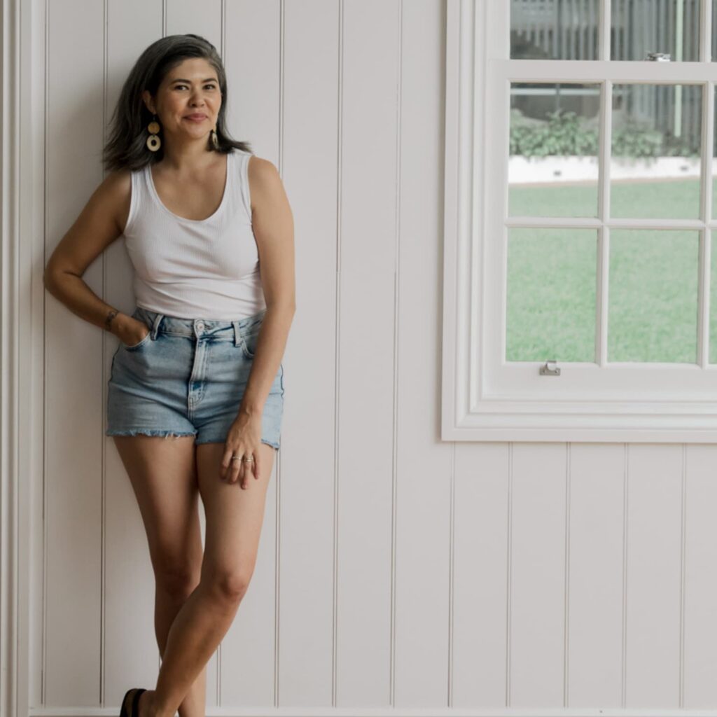An image of a woman wearing a white tank and denim shorts leaning against a white wooden wall.