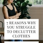 "7 Reasons Why You Struggle to Declutter Clothes + What to Do About It" in a white box with a woman holding a black mug and wearing jeans, a black tank top, and black sandals leaning against a wooden table in the background.
