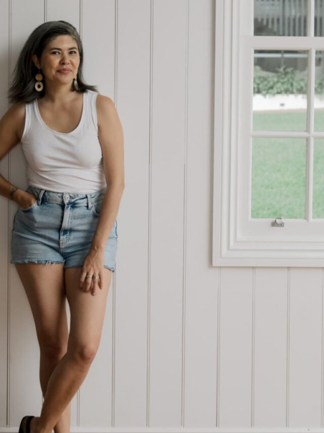 An image of a woman wearing a white tank and denim shorts leaning on a white wooden wall.