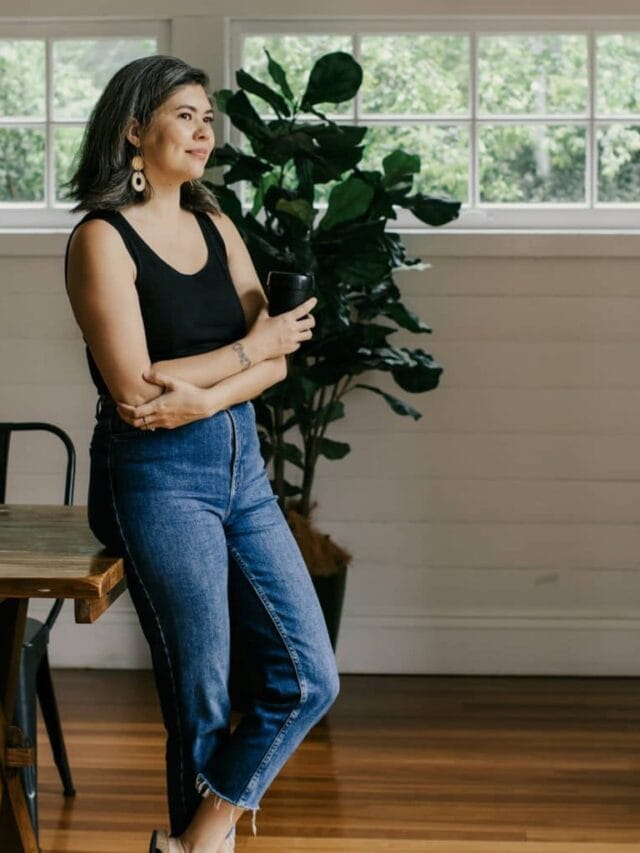 A woman holding a black mug and wearing jeans and a black tank top leaning against a wooden table in a room with white wooden paneled walls and 2 windows.