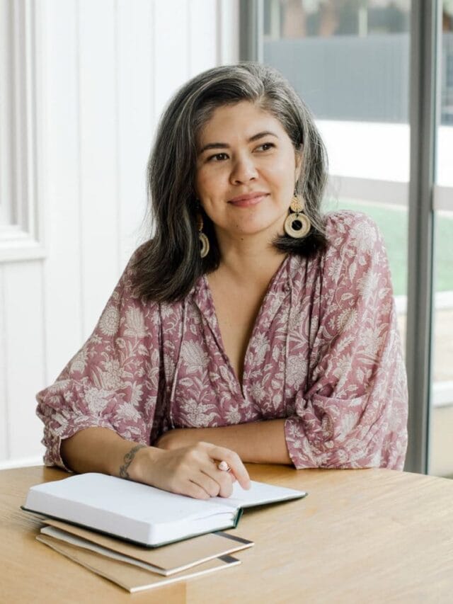 A smiling woman wearing a pale pink with white floral print dress sitting at a wood table holding a pen with an open journal in front of her.