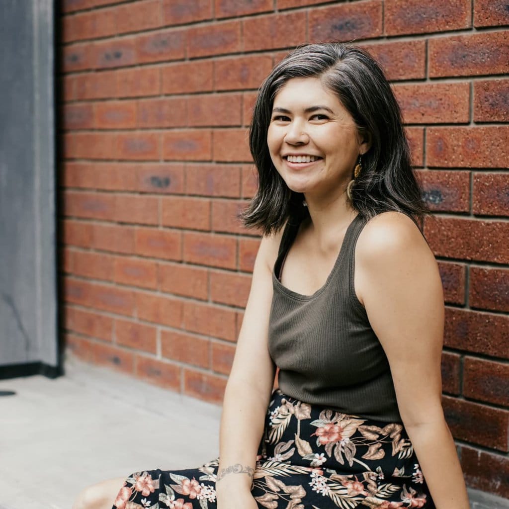 A picture of a woman wearing a grey tank top and black floral skirt leaning on a brick wall and smiling at the camera.