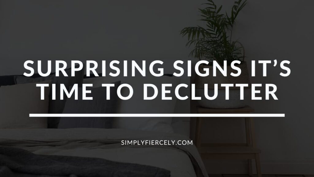 "Surprising Signs It’s Time to Declutter" in white letters on a translucent black overlay on top of an image of a bed with white sheets and a grey blanket, fluffy pillows, and a side table with a plant on it.